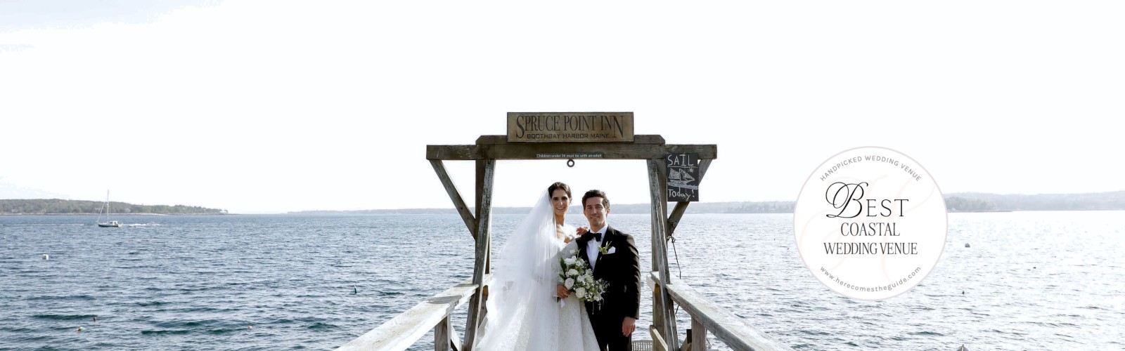 A bride and groom stand on a wooden dock by the water, with small boats visible in the background under a clear sky.