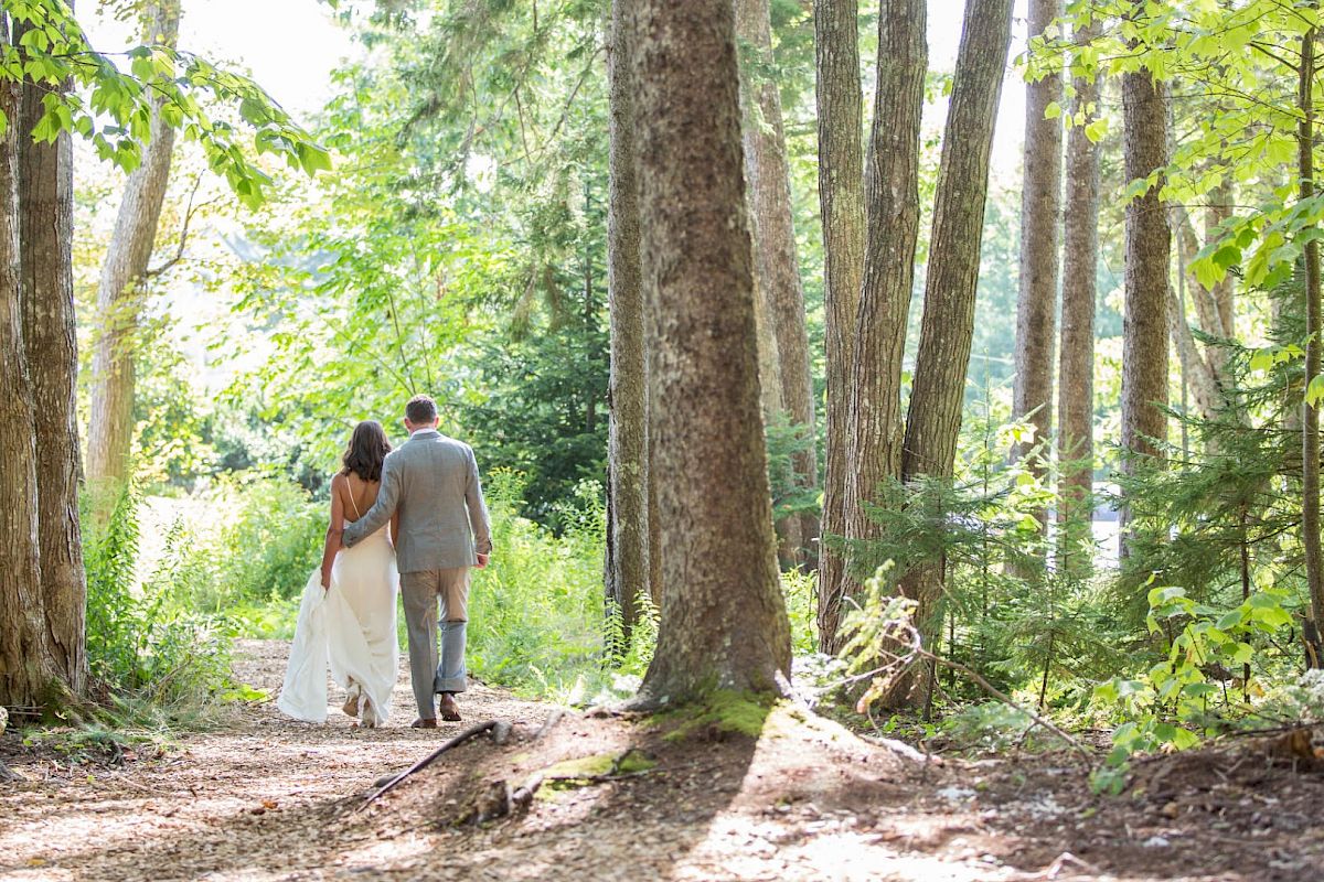 A couple in wedding attire walks hand in hand through a sunlit forest, surrounded by tall trees and lush greenery, heading down a wooded path.