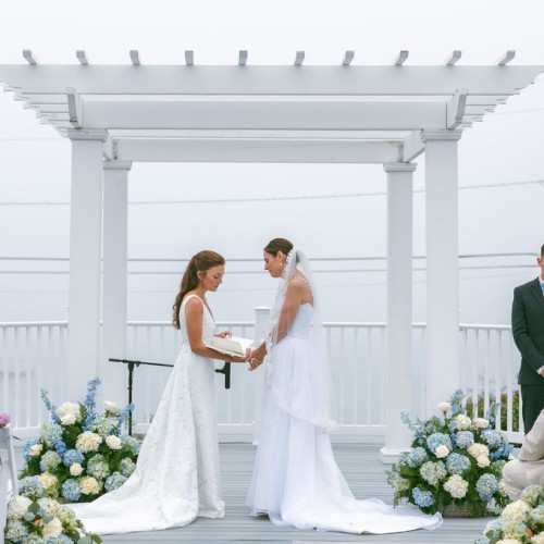 Two brides at an outdoor wedding ceremony under a white pergola, exchanging vows with guests seated nearby, a officiant on the left.