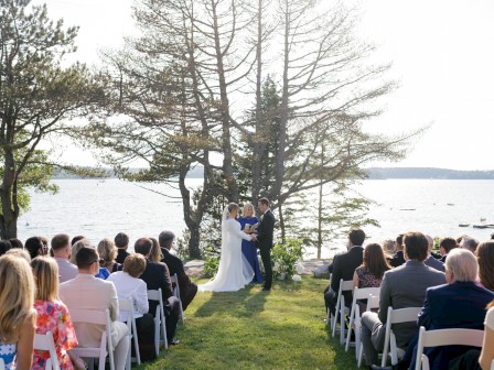 A couple in wedding attire exchange vows beside a lakeside, seated guests on white chairs watch as the ceremony unfolds by a tree backdrop.