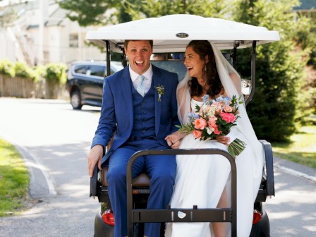 A newlywed couple rides away in a golf cart: the groom in a blue suit, the bride in a white dress holding a bouquet, smiling under a white veil.