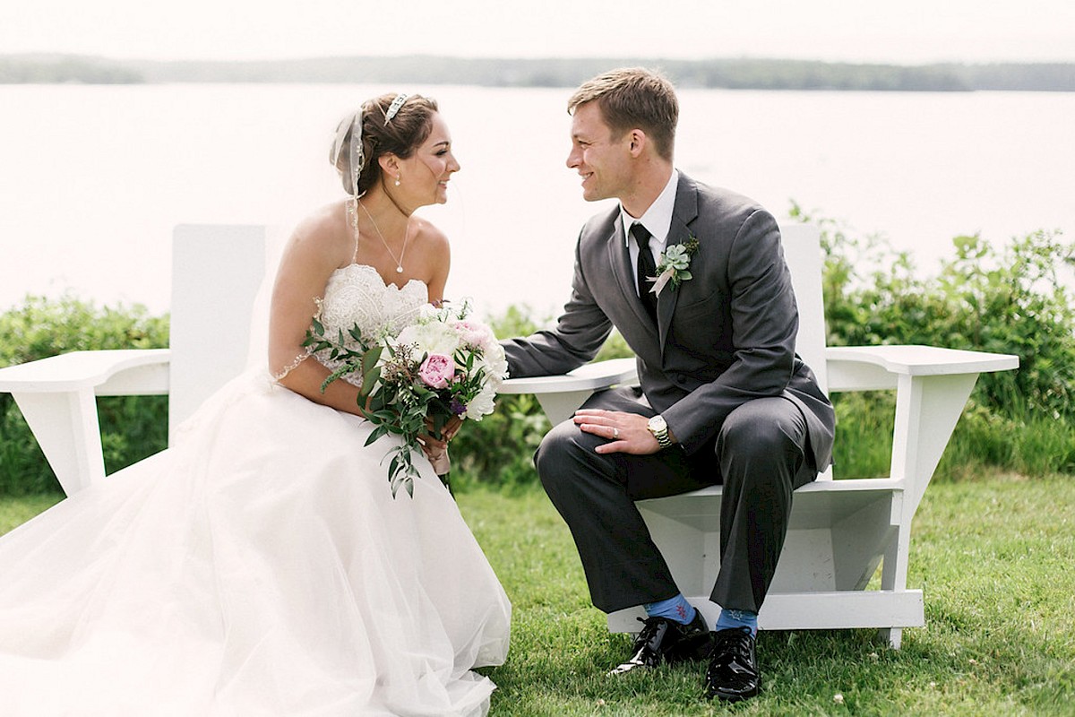 A couple, possibly newlyweds, sits on white benches by a scenic lake, smiling at each other. The bride holds a bouquet of flowers.