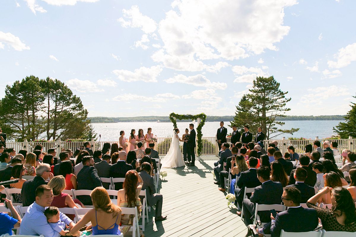 A wedding ceremony is taking place outdoors by a lake, with guests seated and the couple standing under an arch.