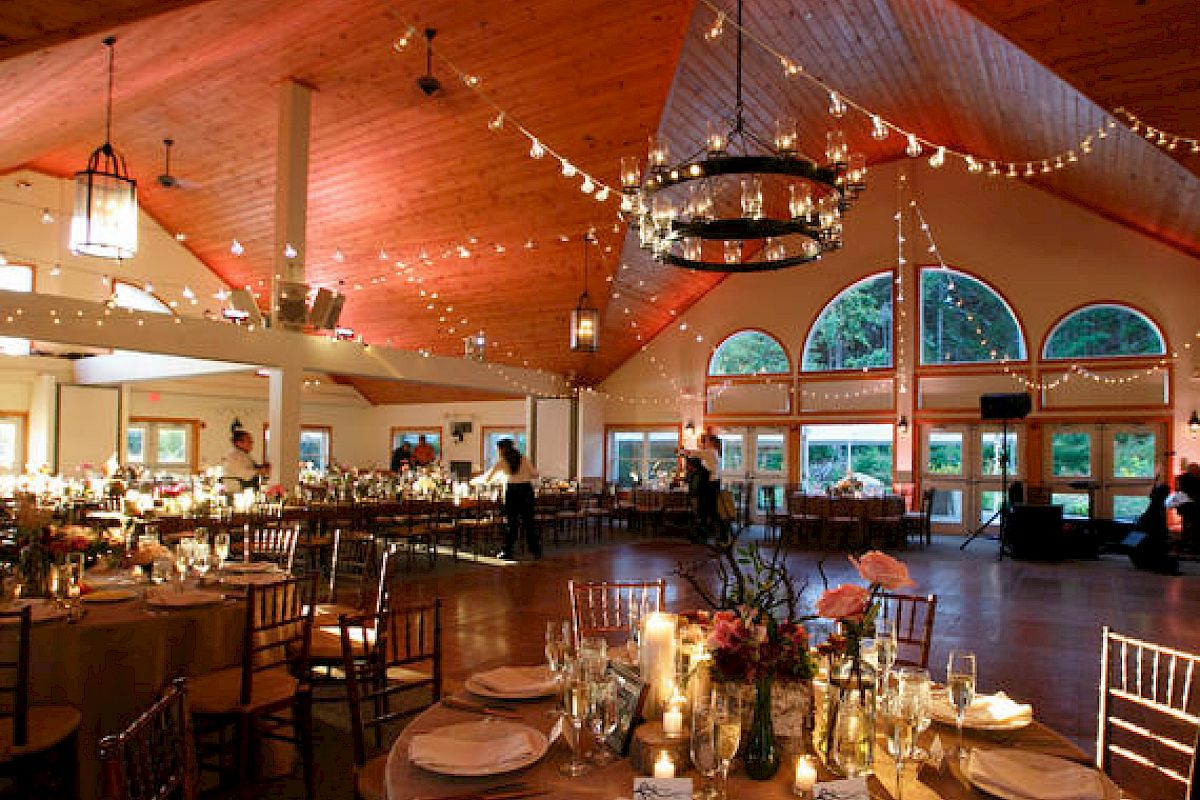 A beautifully decorated event space with round tables set for dining, string lights hanging from the wooden ceiling, and large windows in the background.