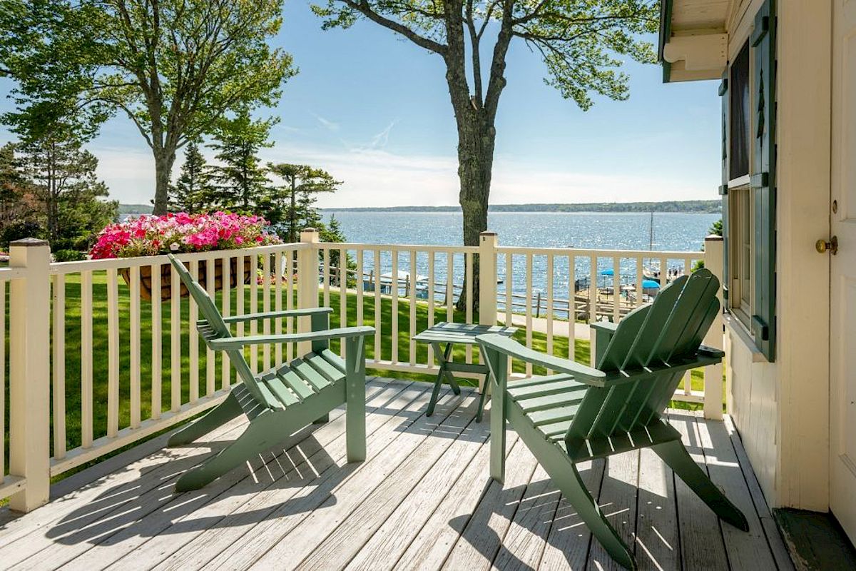 Two green Adirondack chairs on a wooden deck overlook a scenic view of a lake with trees and flowers in the background, under a clear sky.
