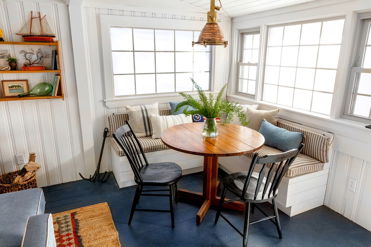 A cozy dining nook with a round wooden table, three chairs, built-in bench seating, a hanging lamp, and a shelf with books and decor items.