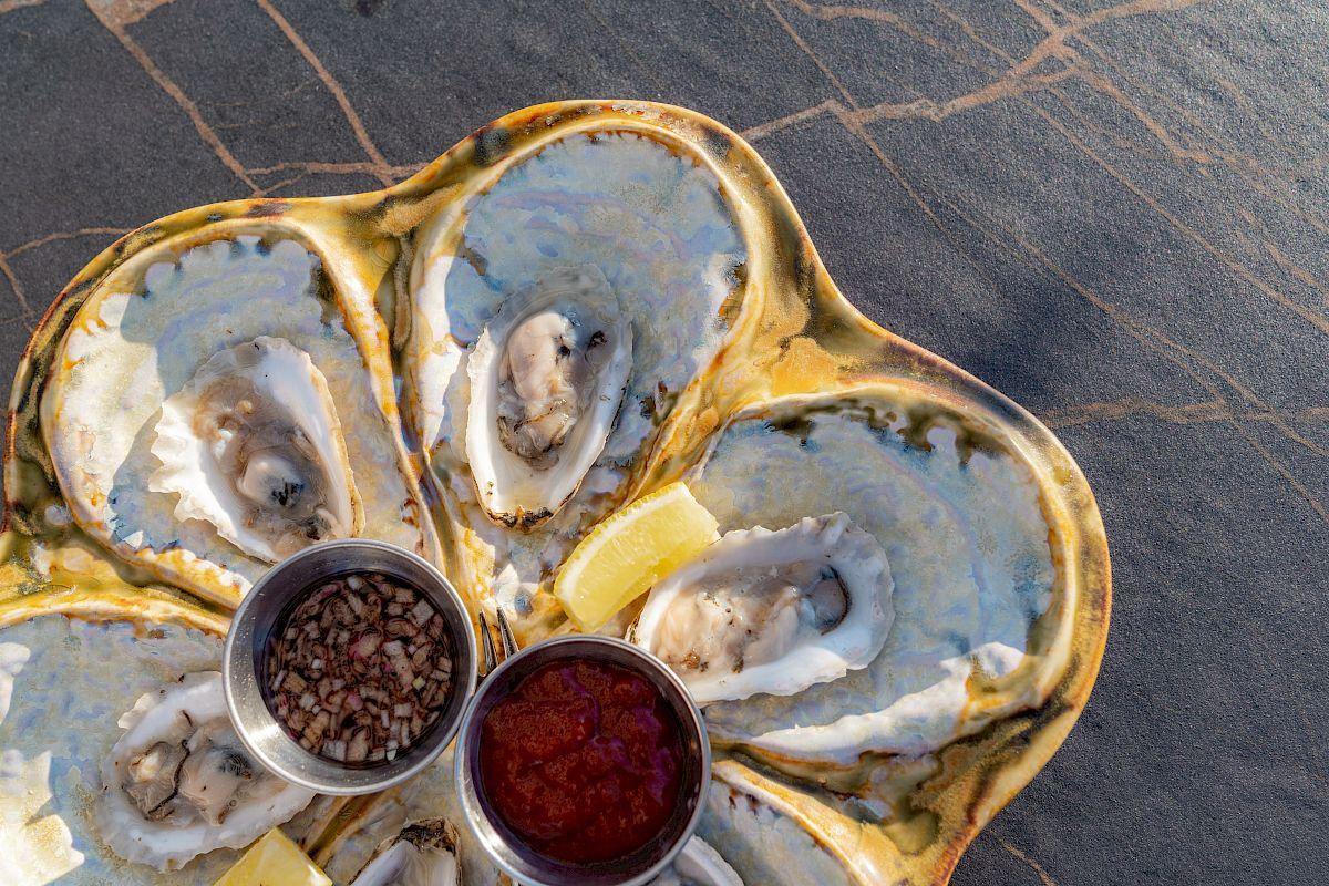 A plate of oysters with two small condiment cups containing toppings and lemon wedges, placed on a dark surface.