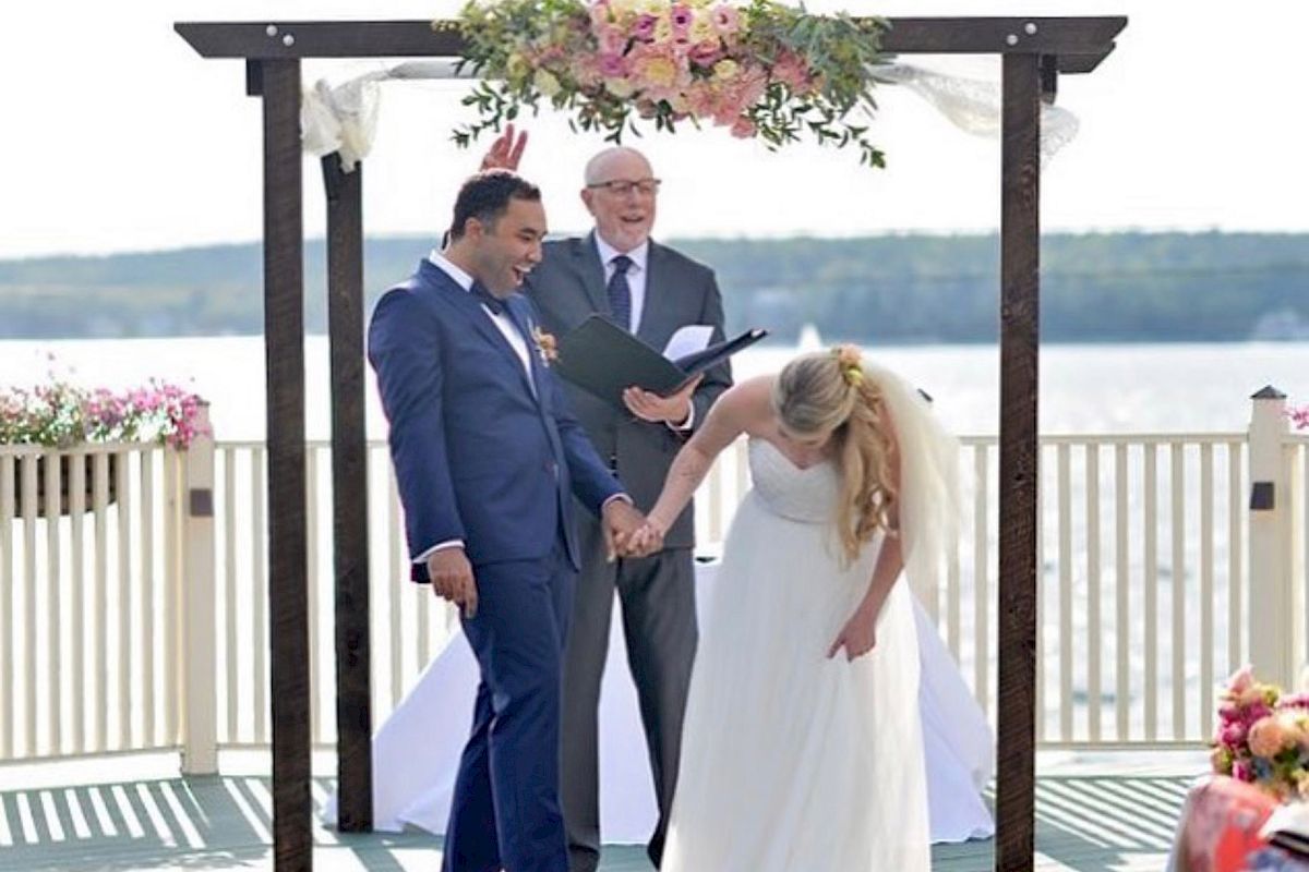 A newlywed couple holds hands and smiles under a floral arch by a lakeside, with an officiant standing behind them, during an outdoor wedding ceremony.