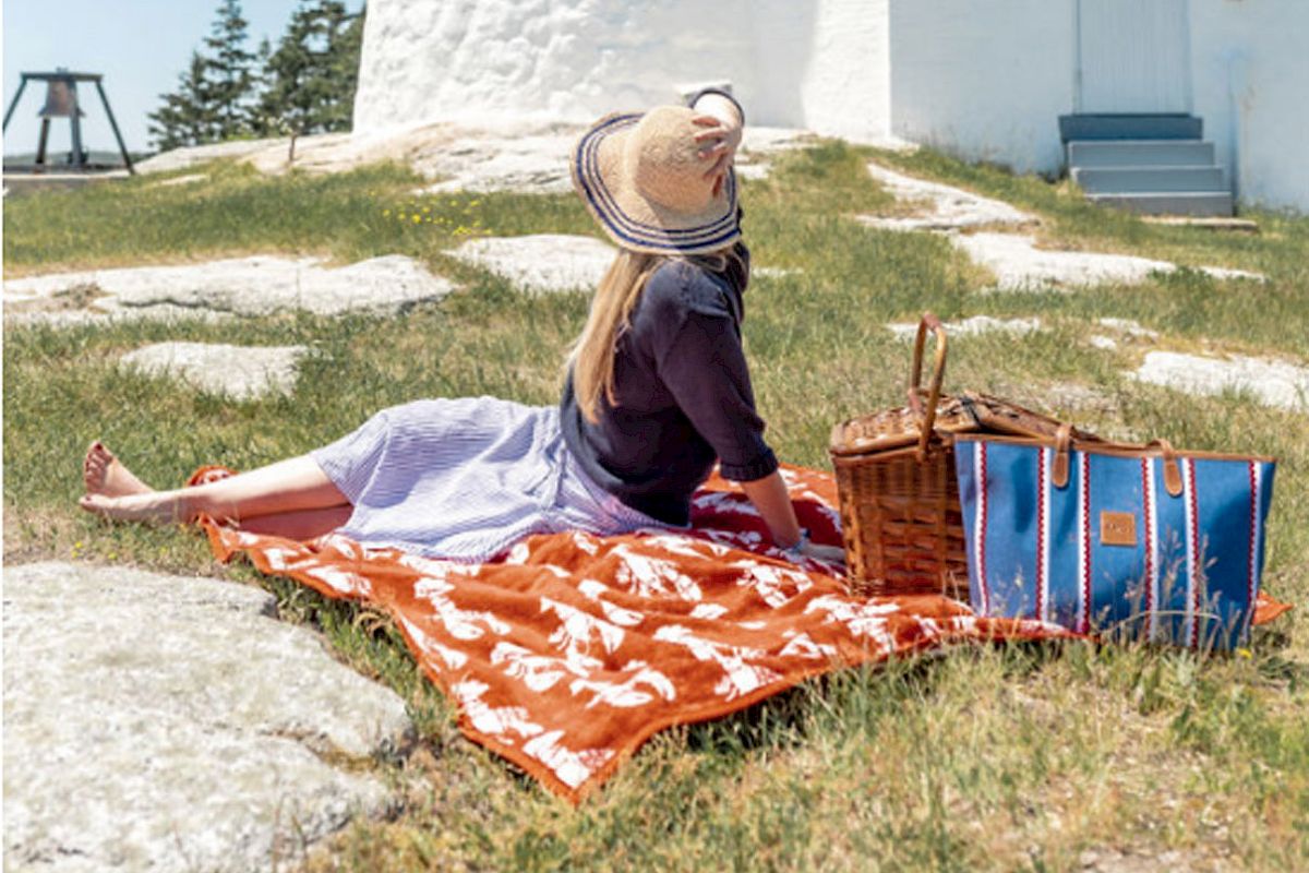 A person sits on a blanket having a picnic near a lighthouse on a bright, sunny day, surrounded by green grass and clear skies.