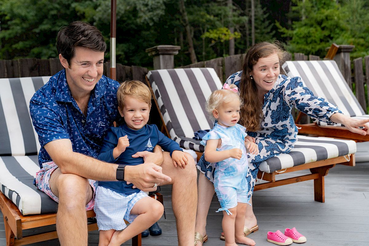 A family of four sits on lounge chairs outdoors; the parents and two young children are smiling, with the mother pointing at something.