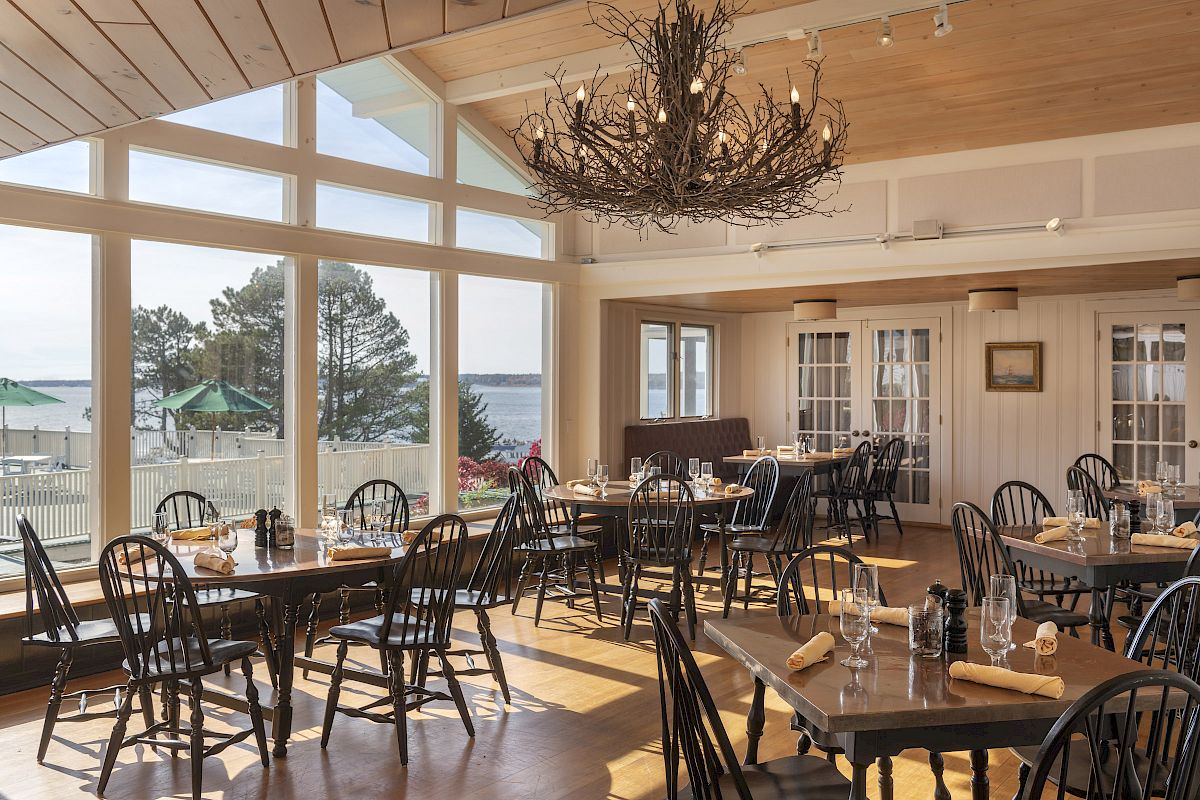 A bright, spacious dining area with wooden tables and chairs, large windows with a scenic outdoor view, and a rustic chandelier hanging from the ceiling.