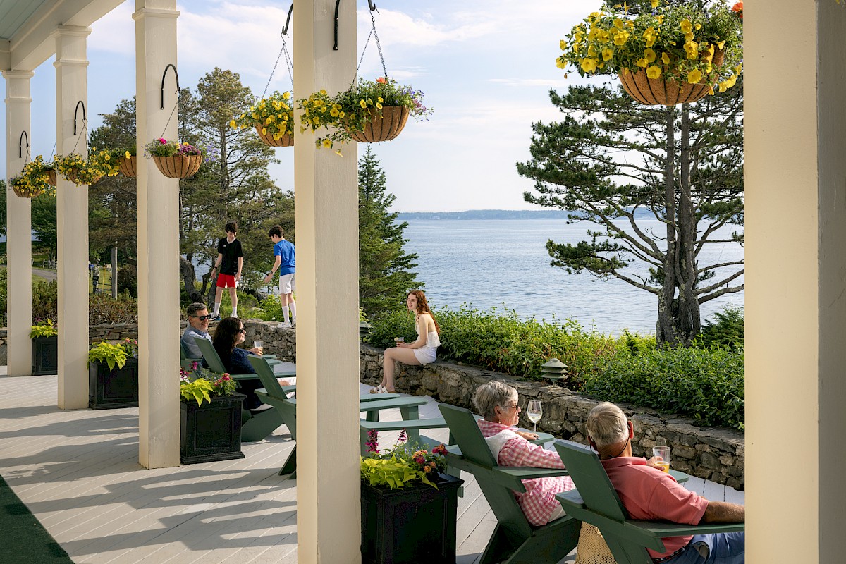 People relaxing on a porch with chairs, hanging flowers, and a scenic ocean view in the background.