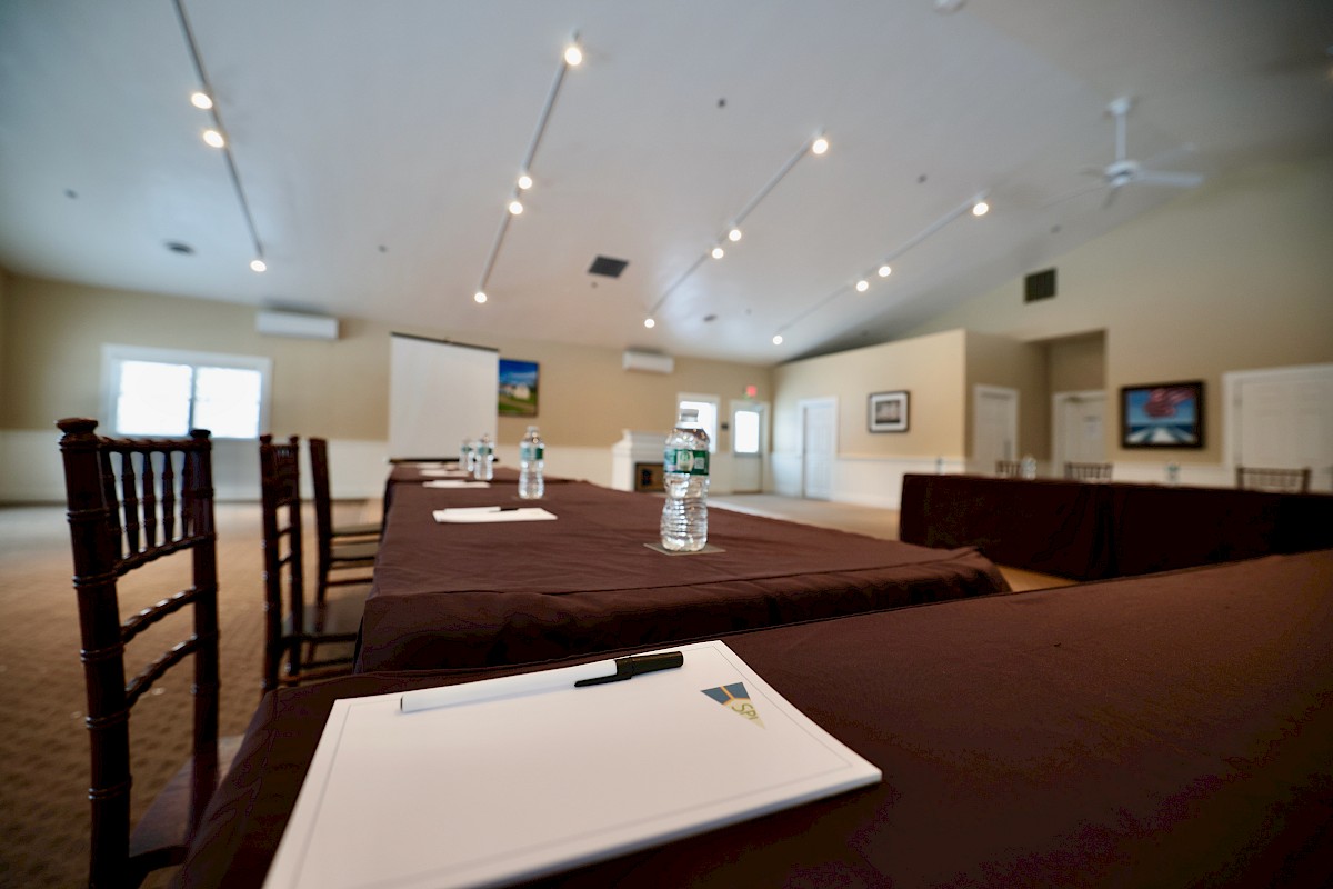 The image shows a conference room with tables covered in brown tablecloths, notepads, pens, and water bottles, facing a presentation screen.