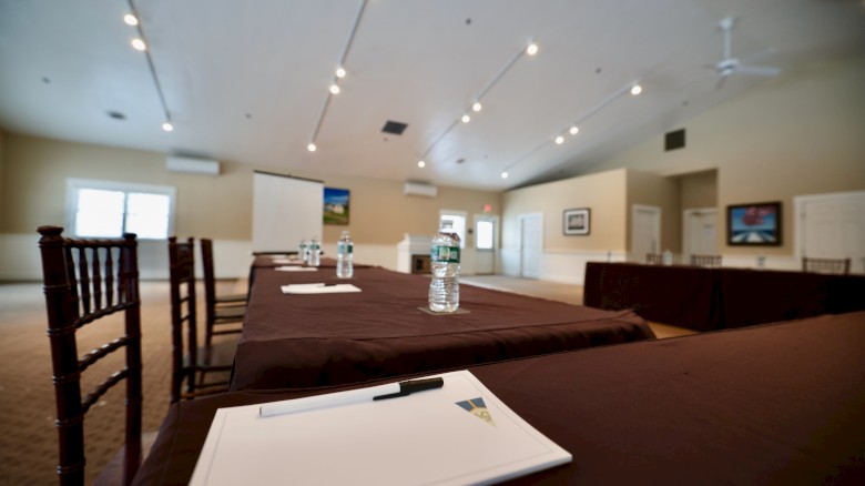 The image shows a conference room with tables covered in brown tablecloths, notepads, pens, and water bottles, facing a presentation screen.