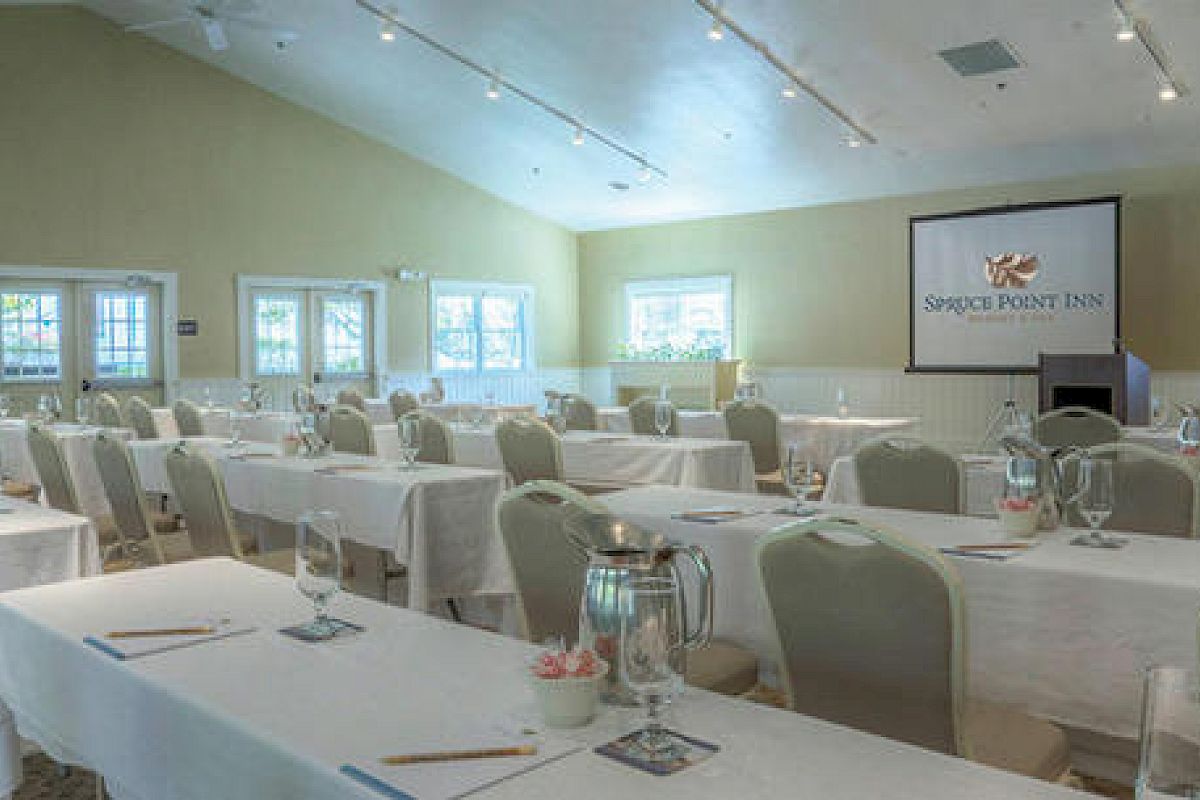 The image shows a conference room set up for a meeting or event with rows of tables and chairs, a screen displaying 