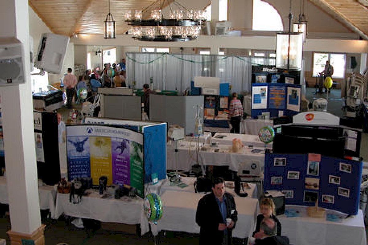 The image shows an indoor exhibition or trade show with various booths, informational displays, attendees, and some decorations like balloons.