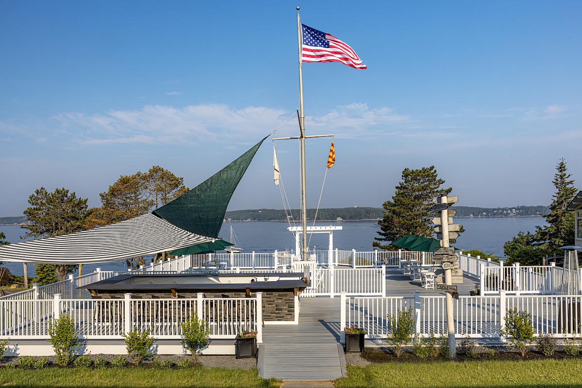 Waterfront area with deck, American flag, sail shade, and plants; view of water and trees in background.