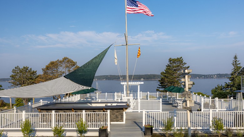 Waterfront area with deck, American flag, sail shade, and plants; view of water and trees in background.