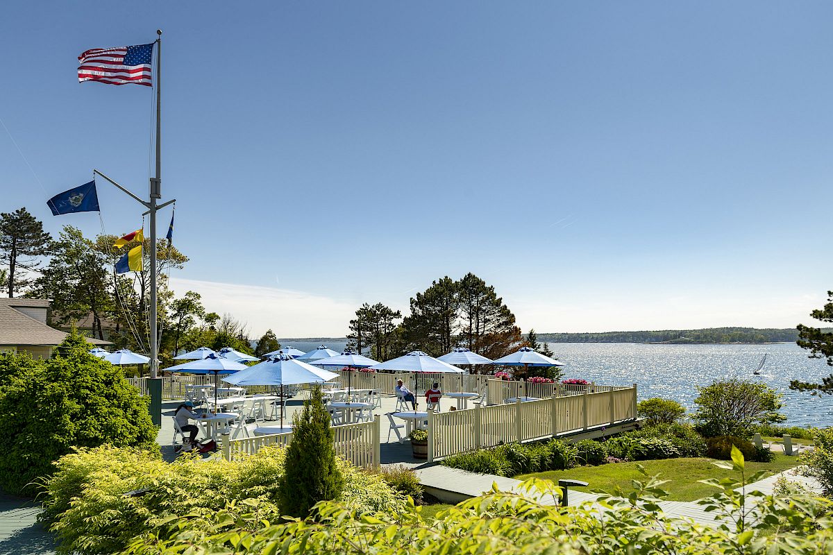 An outdoor seating area by a lake with blue umbrellas, surrounded by lush greenery and flags, all under a clear blue sky.