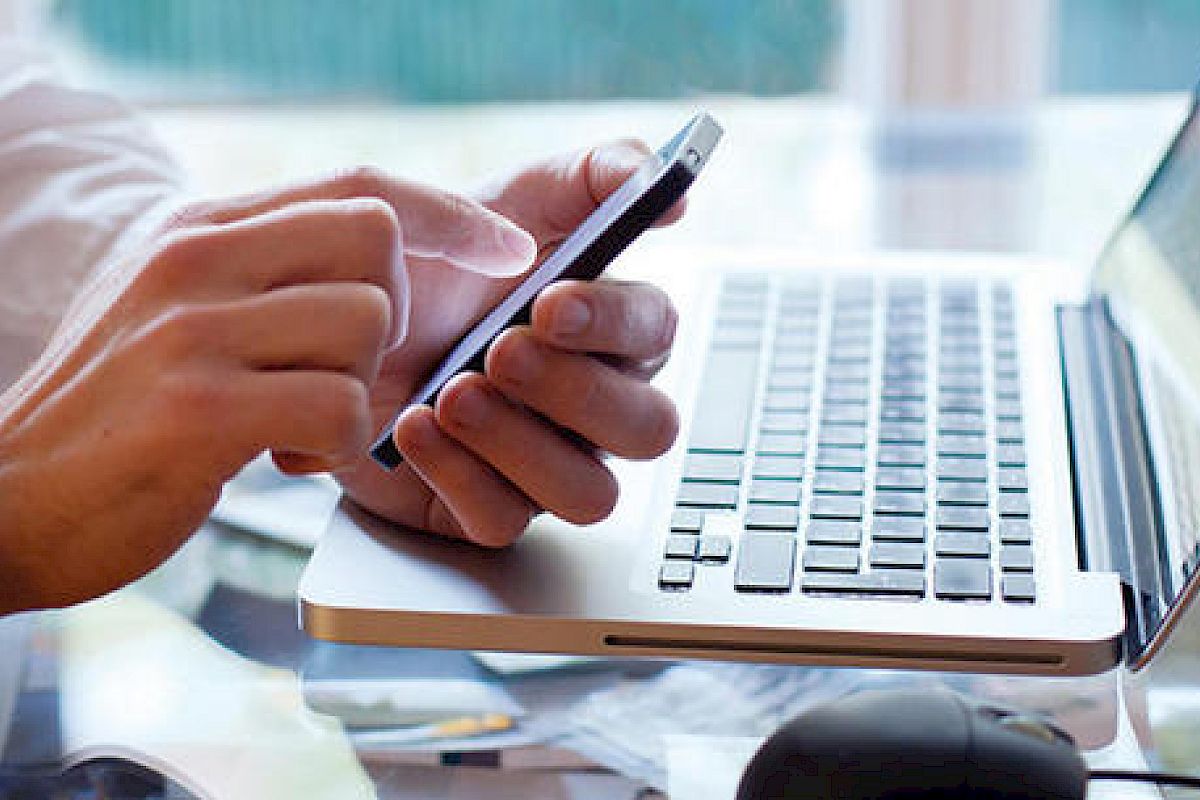 A person is using a smartphone while sitting in front of an open laptop computer on a desk.