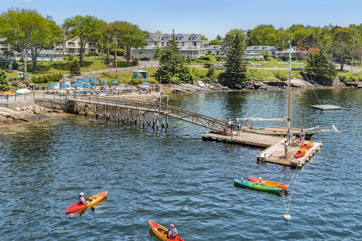 The image shows people kayaking near a wooden dock with a small sailboat, located by a waterfront community with houses and greenery in the background.