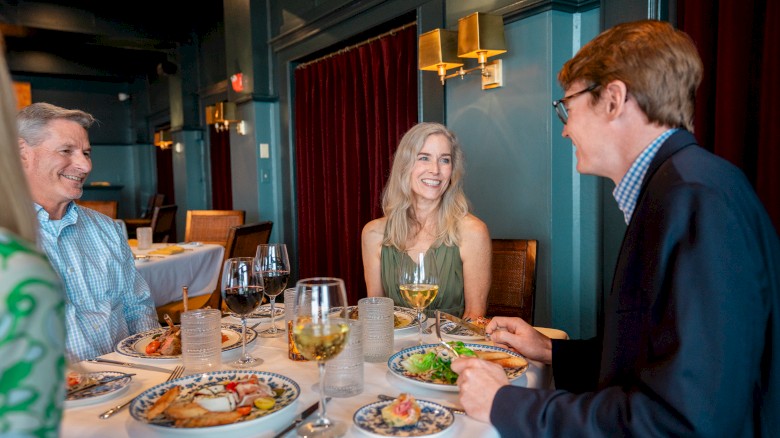 Three people are dining at a restaurant, smiling and enjoying food and drinks at a table set with plates and glasses.
