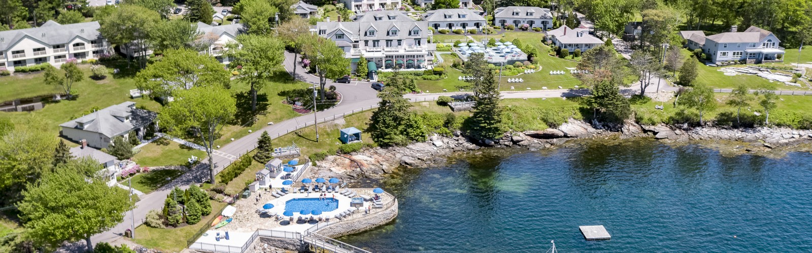 Aerial view of a waterfront resort with multiple buildings, a dock, boats, and a swimming pool, surrounded by lush greenery and water.