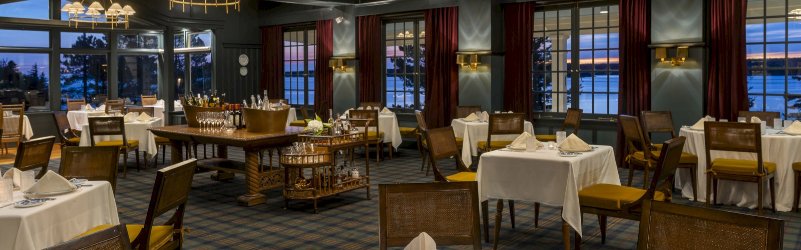 A dimly lit, elegant restaurant with white tablecloths, folded napkins, and fine tableware, featuring large windows and hanging light fixtures.