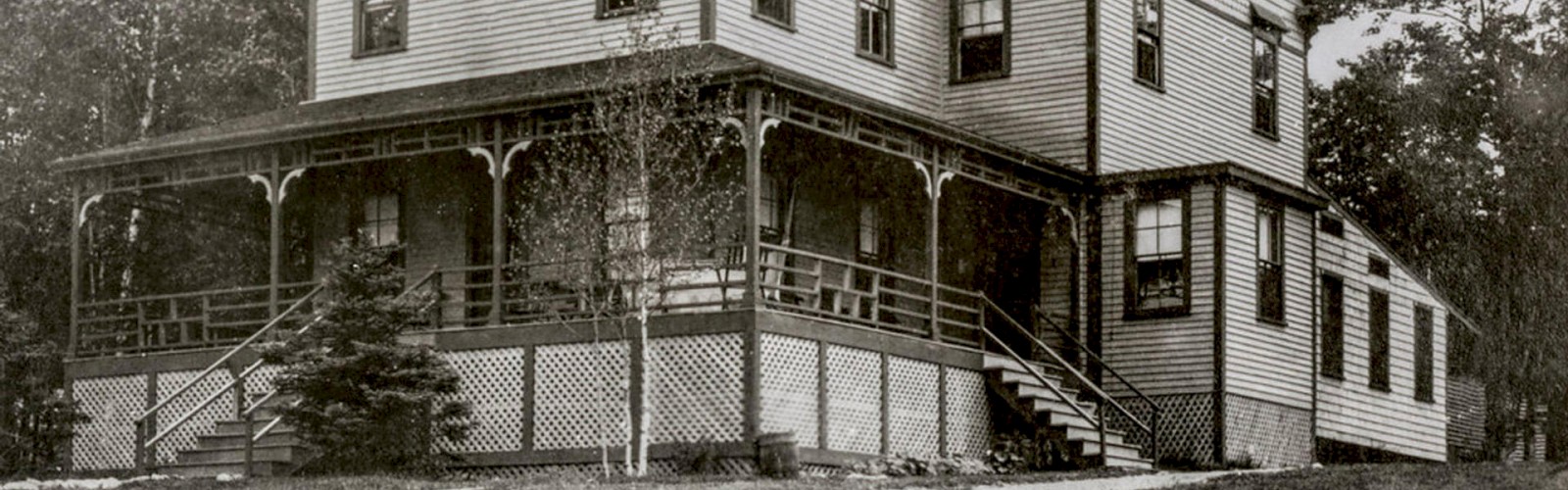 An old, black-and-white photo of a large, two-story house with a wraparound porch, multiple windows, and surrounding trees.
