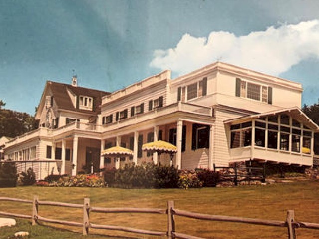 The image shows a large house with a sloped roof, multiple windows, a porch, and a grassy area in front, enclosed by a wooden fence. Ending the sentence.