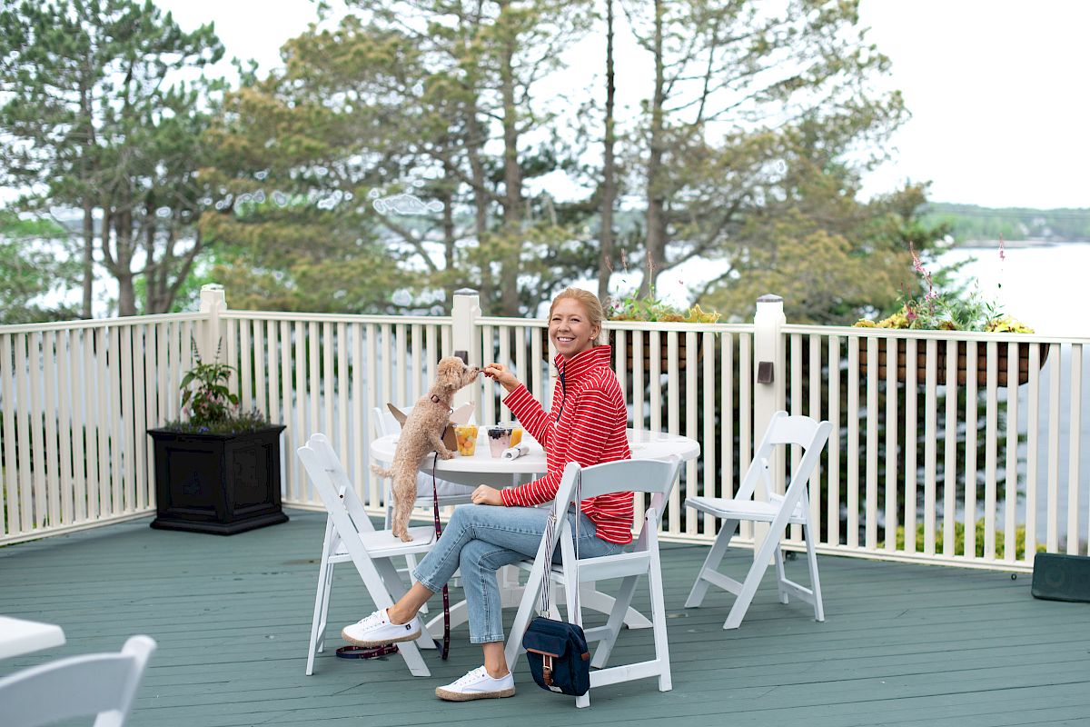 A woman in a red striped sweater sits at an outdoor table with her dog on a deck, surrounded by trees and overlooking a body of water.