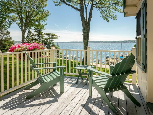 A cozy deck with two green Adirondack chairs overlooks a serene lake, surrounded by trees and colorful flowers, under a bright blue sky.