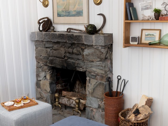 A cozy living room with a stone fireplace, a blue ottoman, wall shelves with decor, a painting above the fireplace, and a basket with firewood.