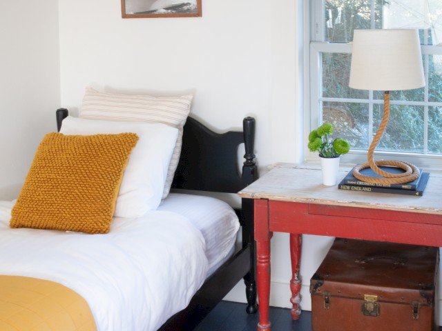A cozy bedroom with a single bed, mustard pillow and throw, red side table with a lamp, plant, and trunk, featuring nautical decor and a window.