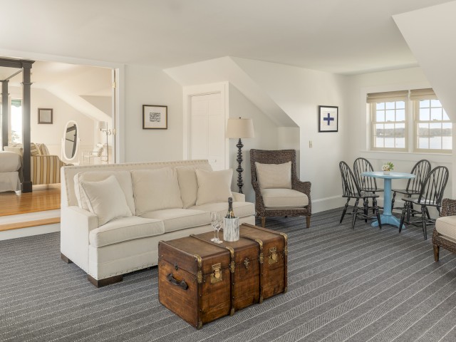 A cozy living room features a beige sofa, vintage trunk coffee table, wicker chair, and small dining area with a table and four chairs.