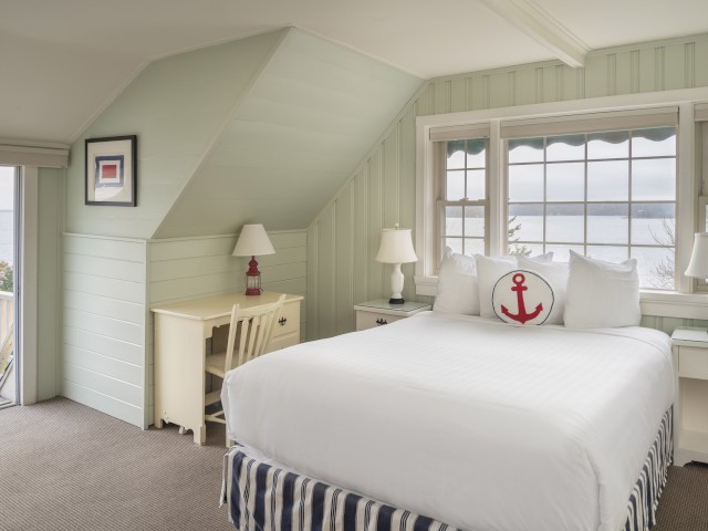 A cozy bedroom with nautical decor, featuring a bed, a desk, two nightstands with lamps, and large windows with an ocean view.
