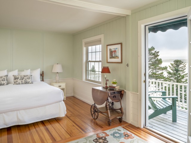 A cozy bedroom with a white bed, side table, lamp, and wooden floors opens to a balcony with a green chair and scenic outdoor view.