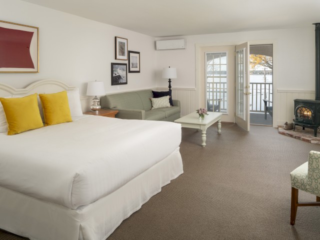 A spacious, well-lit room with a bed, sofa, tables, and a fireplace, opening to a balcony. Decor includes framed art and cushioned chairs.