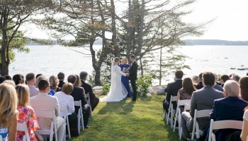 A wedding ceremony outdoors by a lake, couple kissing under trees while guests sit in white chairs.