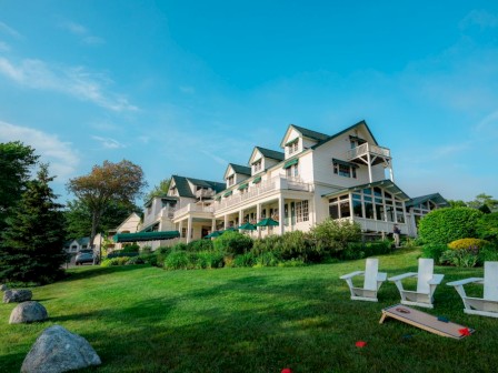 A large resort-style house with a green lawn, white chairs by the yard, trees, and a bright blue sky.