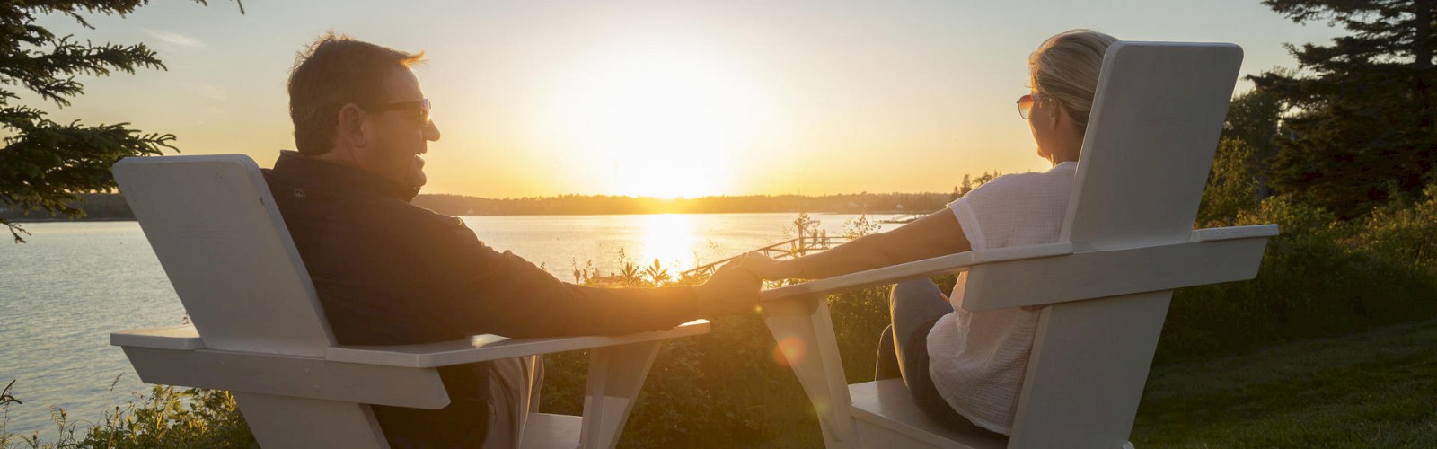 A couple sits in white Adirondack chairs, holding hands and watching the sunset by a lake, surrounded by trees and green grass.
