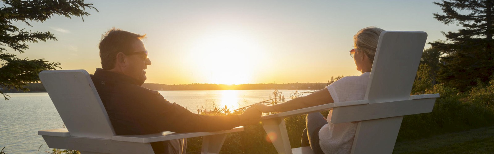 Two people are seated in Adirondack chairs, holding hands, and enjoying a sunset by a lakeside, surrounded by trees and nature.