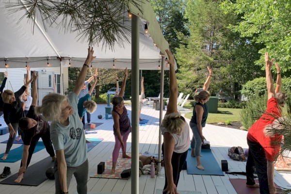 A group of people practicing yoga outdoors on mats, surrounded by trees and a tent.