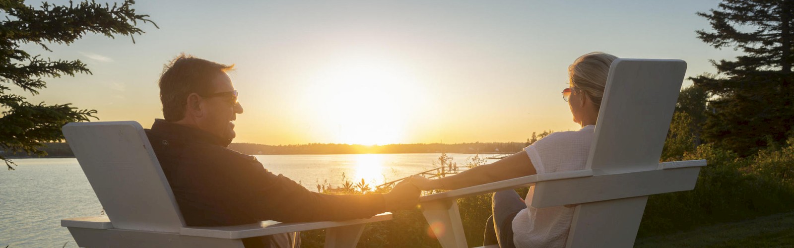 Two people are sitting in Adirondack chairs, holding hands, and watching the sunset over a lake, surrounded by trees and greenery.