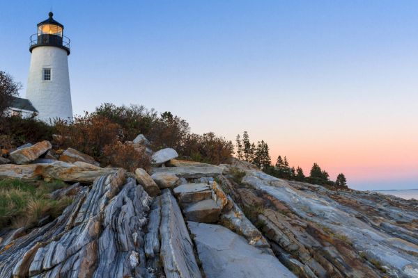 A lighthouse on a rocky shoreline during sunset, with a partly cloudy sky and surrounding trees ending the sentence.