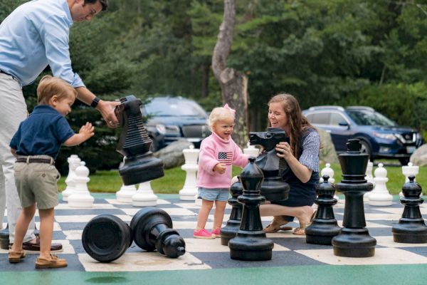 A family enjoys a game on a giant outdoor chessboard, with a man and two kids holding large chess pieces while a woman engages with a small child.