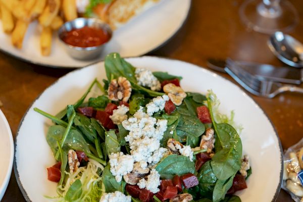 A fresh salad with spinach, cheese, nuts, and beets is served on a white plate. In the background, there are fries with ketchup.