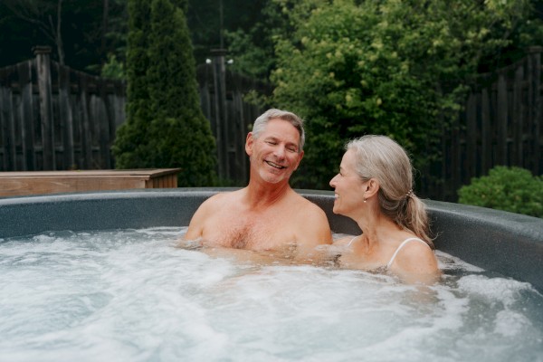 A couple is relaxing and smiling together in an outdoor hot tub, surrounded by greenery in the background.