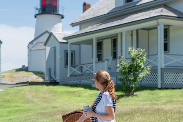 A person is sitting on a picnic blanket near a house with a lighthouse in the background on a sunny day.