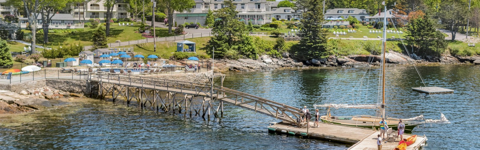 A scenic waterfront with a dock, kayaks, and a sailboat moored at it. There are buildings and trees in the background, with people on the dock.